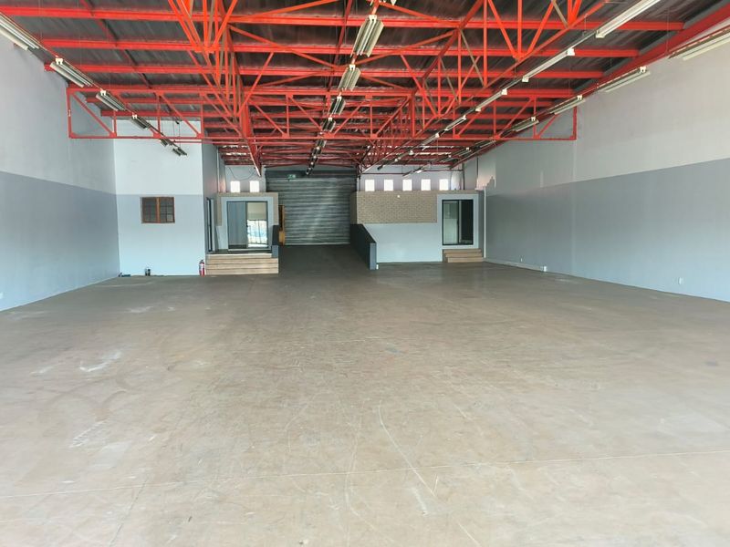 Spacious open-plan retail or industrial warehouse unit featuring high exposed steel roof trusses, concrete flooring, and raised office areas with a roller shutter door.