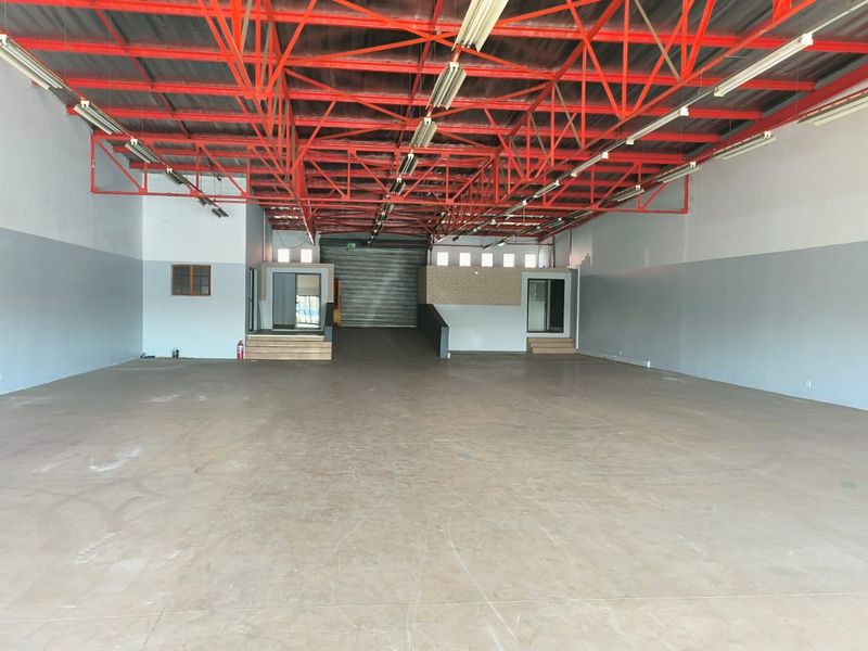 Spacious open-plan industrial or retail utility space featuring high exposed steel roof trusses, concrete flooring, and a raised office area with roller shutter access.