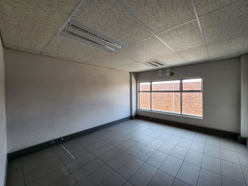 An empty office space featuring tiled flooring, a suspended ceiling with recessed lighting, and large windows providing natural light.