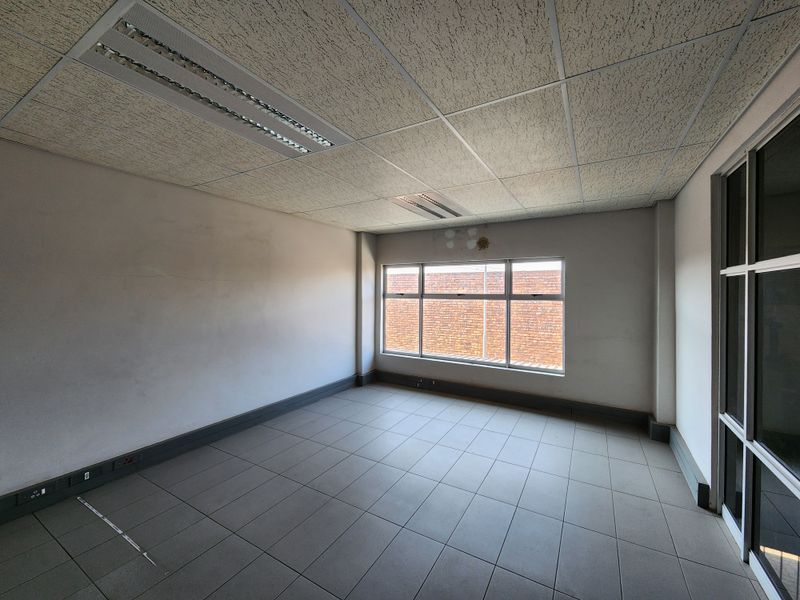 An empty office space featuring tiled flooring, a suspended ceiling with recessed lighting, and large windows providing natural light.