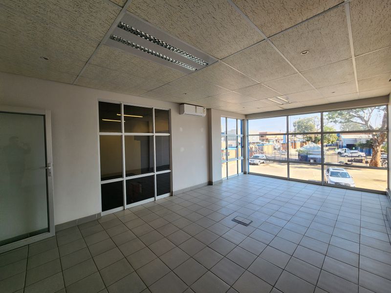 A bright, open-plan office space featuring tiled flooring, large windows providing natural light, a suspended ceiling, and a wall-mounted air conditioning unit.