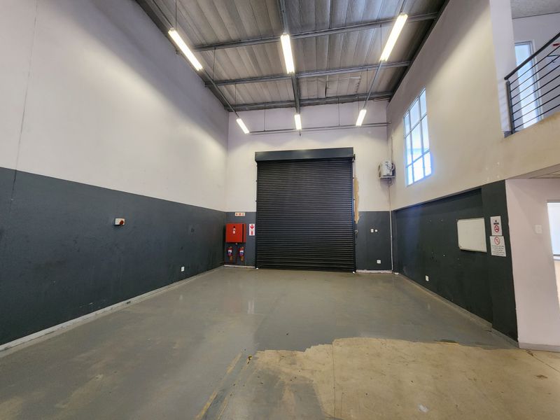 Spacious industrial garage featuring a high roller shutter door, exposed steel roof structure, mezzanine level, and functional fire safety equipment.
