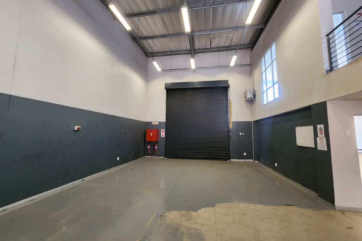 Spacious industrial garage featuring a high roller shutter door, exposed steel roof structure, mezzanine level, and functional fire safety equipment.