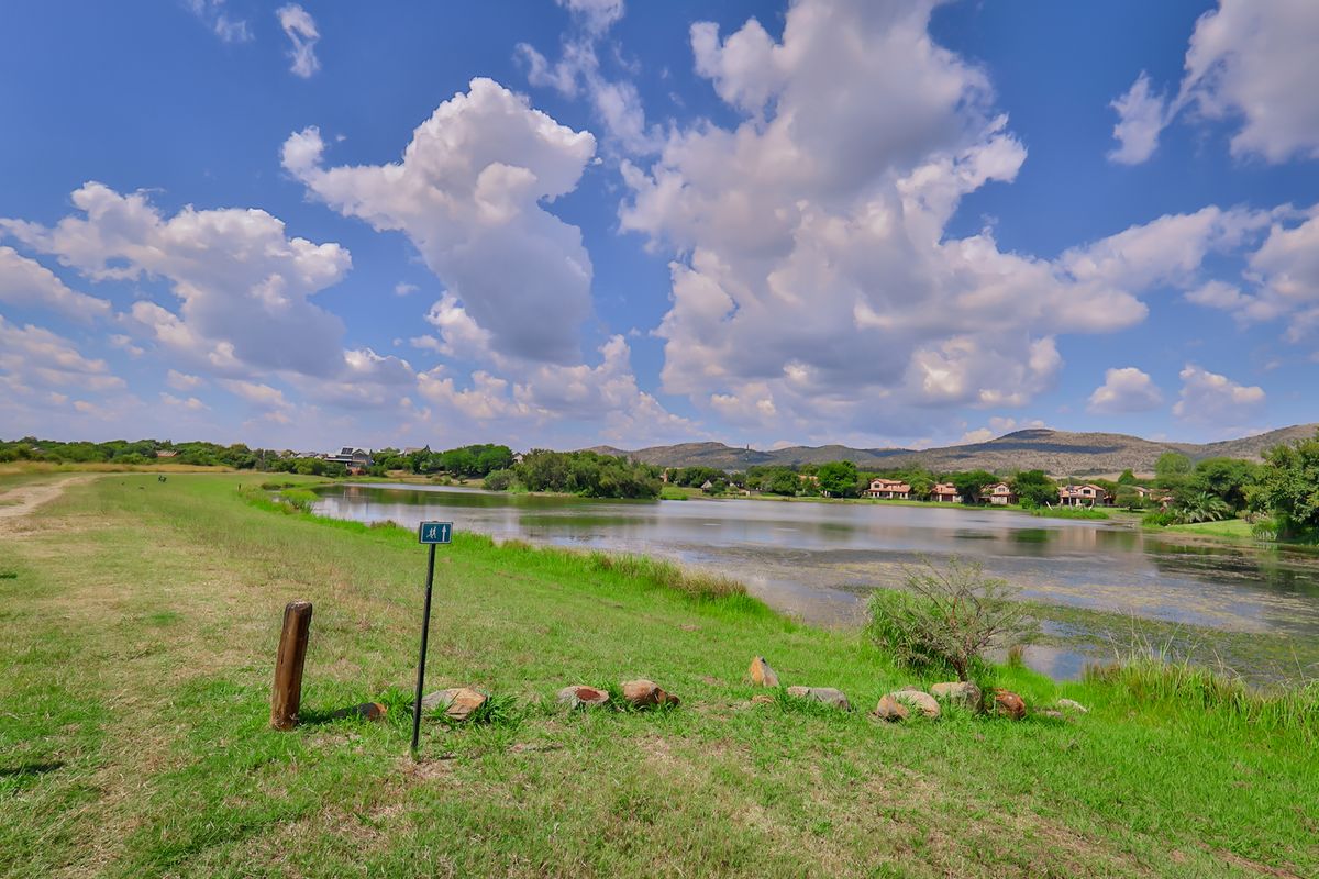 A scenic view of a tranquil lake surrounded by lush greenery and distant residential properties under a bright, cloudy sky.