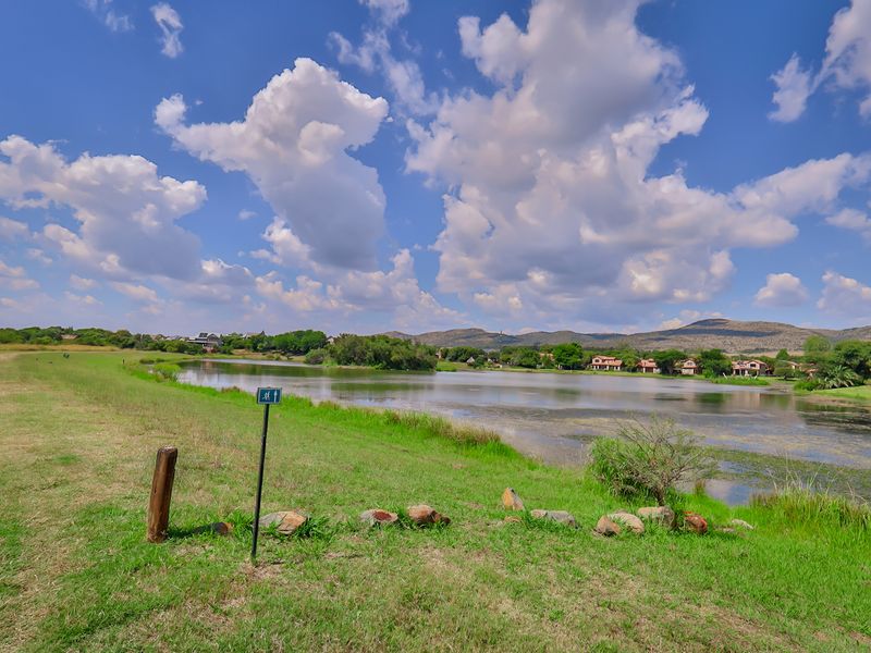A scenic view of a tranquil lake surrounded by lush greenery and distant residential properties under a bright, cloudy sky.