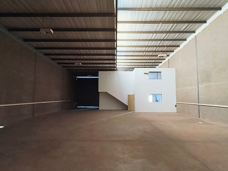 Spacious industrial warehouse featuring high ceilings with exposed steel trusses, translucent roof panels for natural light, concrete flooring, and an integrated mezzanine office structure.