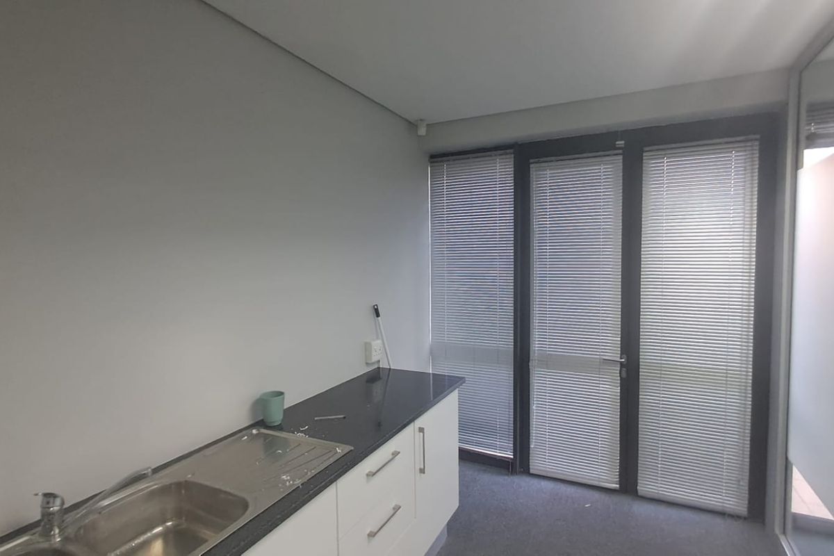 A compact office kitchenette featuring a stainless steel sink, dark laminate countertop, white base cabinetry, and large windows with venetian blinds.
