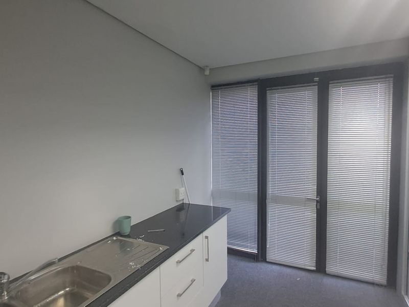 A compact office kitchenette featuring a stainless steel sink, dark laminate countertop, white base cabinetry, and large windows with venetian blinds.