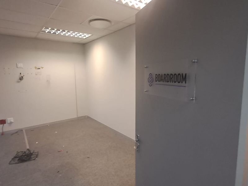 An empty office space designated as a boardroom, featuring a suspended ceiling, recessed lighting, and carpeted flooring, currently requiring minor wall repairs.