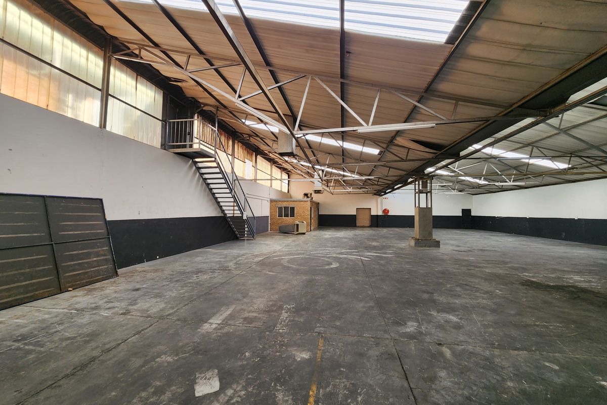 Spacious industrial warehouse featuring high ceilings with exposed steel trusses, translucent roof panels for natural light, concrete flooring, and a metal staircase to mezzanine.