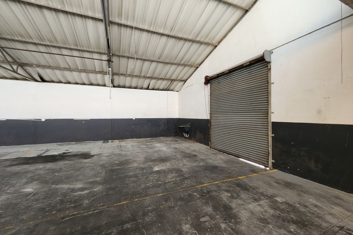 Spacious industrial utility area featuring high ceilings, concrete flooring, a large roller shutter door, and a wall-mounted basin for functional use.