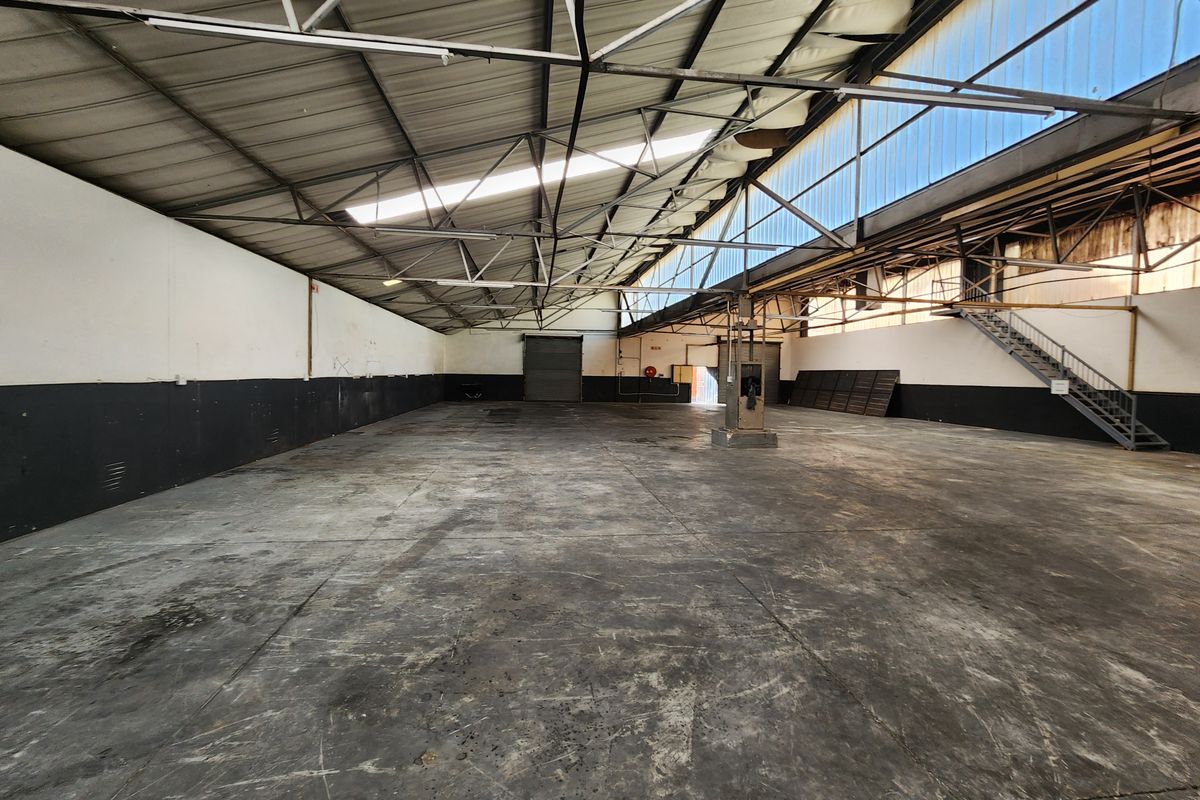 Spacious industrial warehouse featuring high ceilings with exposed steel trusses, concrete flooring, a roller shutter door, and an internal metal staircase.