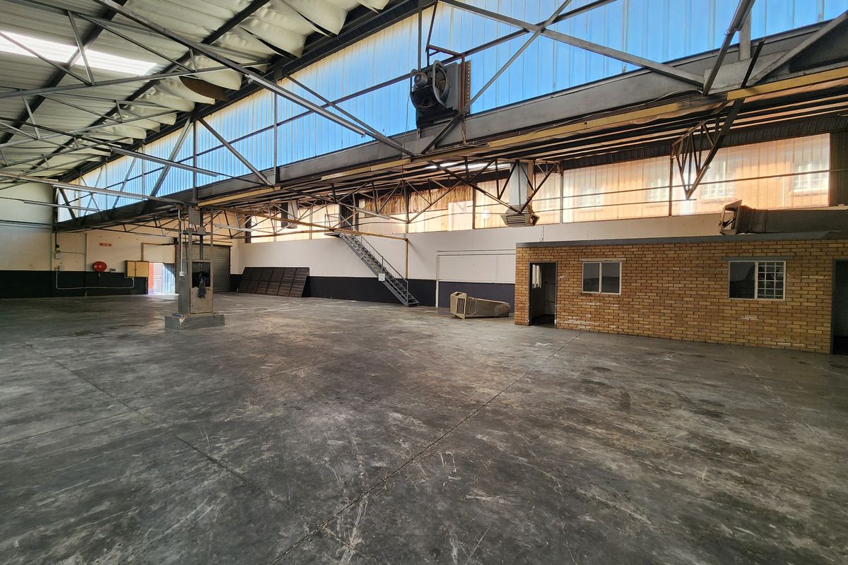 Spacious industrial warehouse featuring high exposed steel truss ceilings, translucent roof panels for natural light, concrete flooring, and an integrated brick office structure.