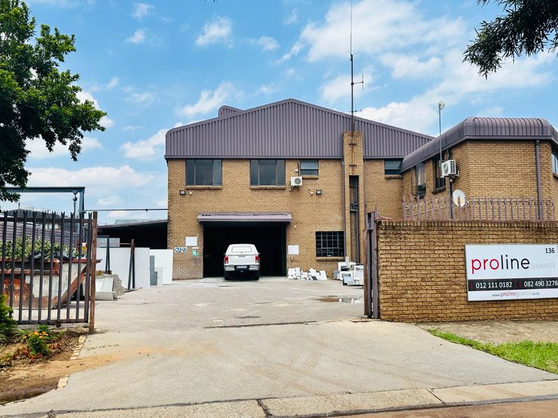 Front exterior view of an industrial property featuring a brick facade, metal roofing, secure perimeter fencing, and a paved driveway entrance.