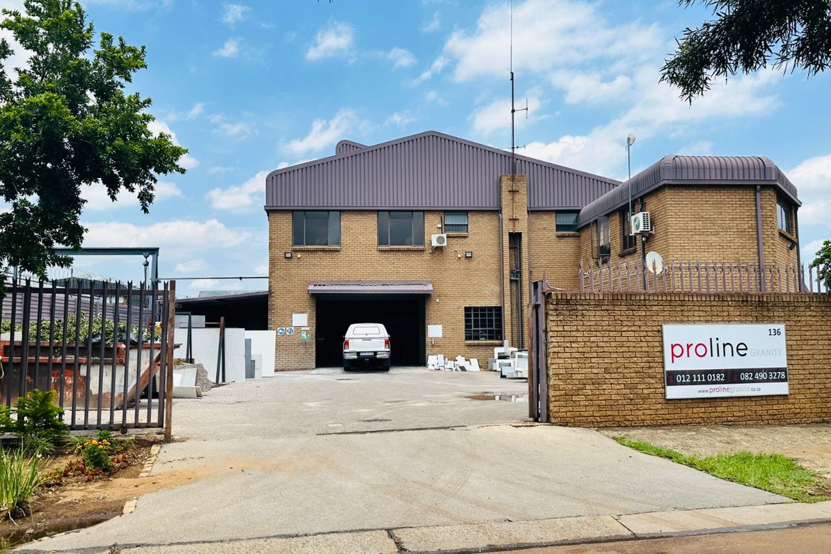 Front exterior view of an industrial property featuring a brick facade, metal roofing, secure perimeter fencing, and a paved driveway entrance.