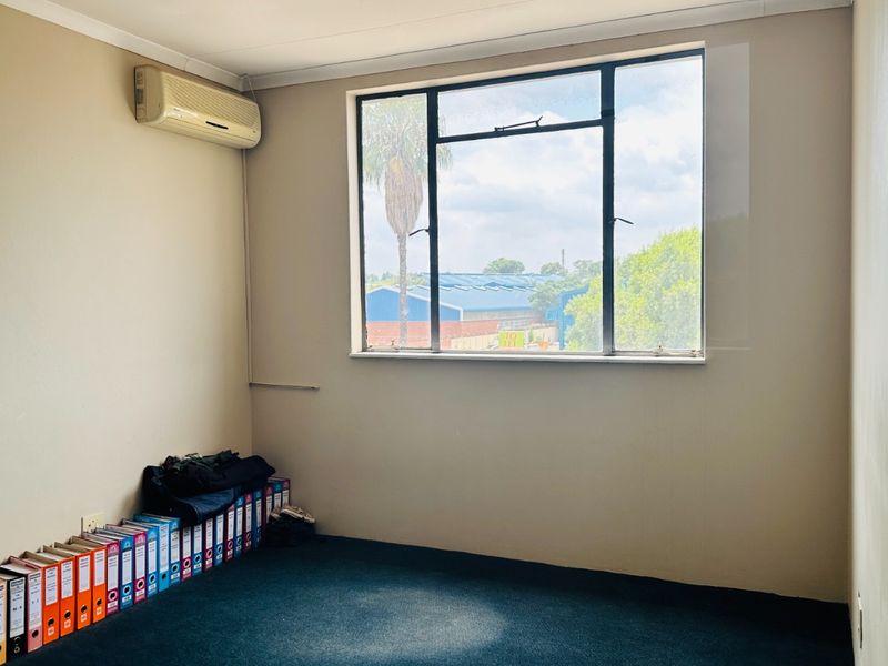 A functional office space featuring carpeted flooring, a split-system air conditioner, and a large window providing natural light and an industrial view.
