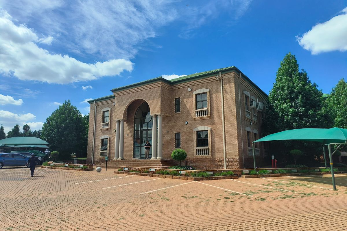 A professional office building featuring a classic brick facade, arched entrance with columns, and an extensive paved parking area with shade cloth carports.