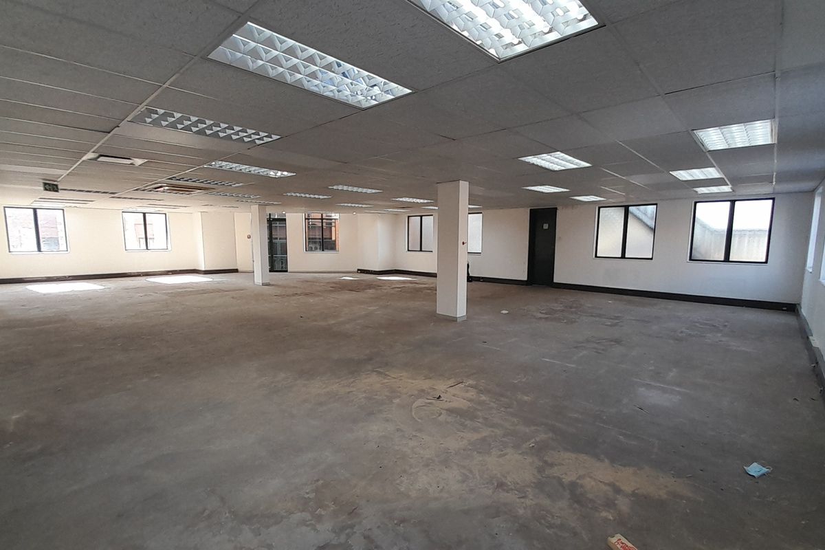 A large, open-plan office space featuring a suspended ceiling with recessed lighting, structural columns, and multiple windows providing natural light.