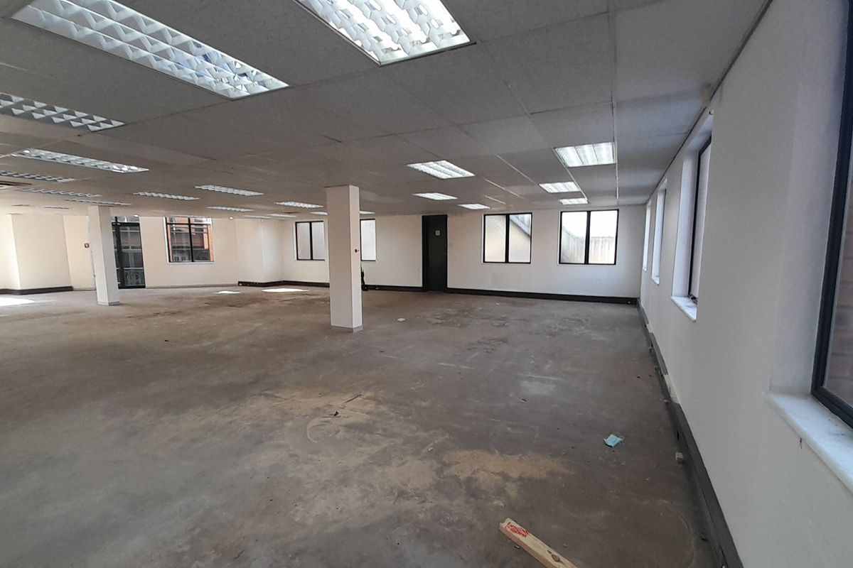A large, open-plan office space featuring a suspended ceiling with recessed lighting, structural columns, and expansive windows providing natural light.