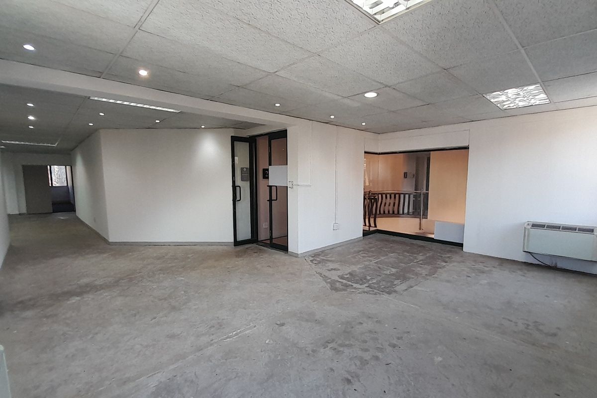 An empty, open-plan office space featuring a suspended ceiling, recessed lighting, and a glass partition door, with concrete flooring throughout.