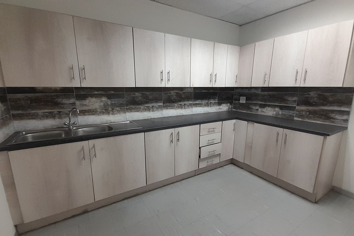 A functional office kitchenette featuring light wood-finish cabinetry, dark countertops, a stainless steel sink, and a decorative tiled backsplash.