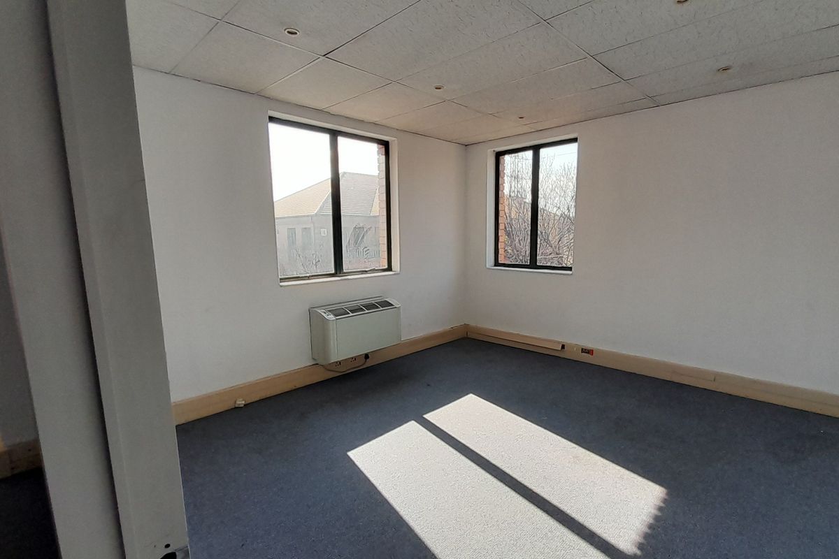 A vacant office space featuring a suspended ceiling, recessed lighting, wall-mounted HVAC unit, and carpeted flooring with natural light from two windows.