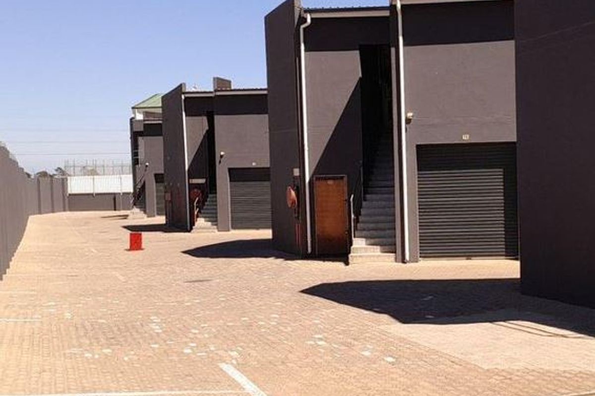 Exterior view of an industrial complex featuring multiple units with roller shutter doors, external staircases, and a paved driveway area.