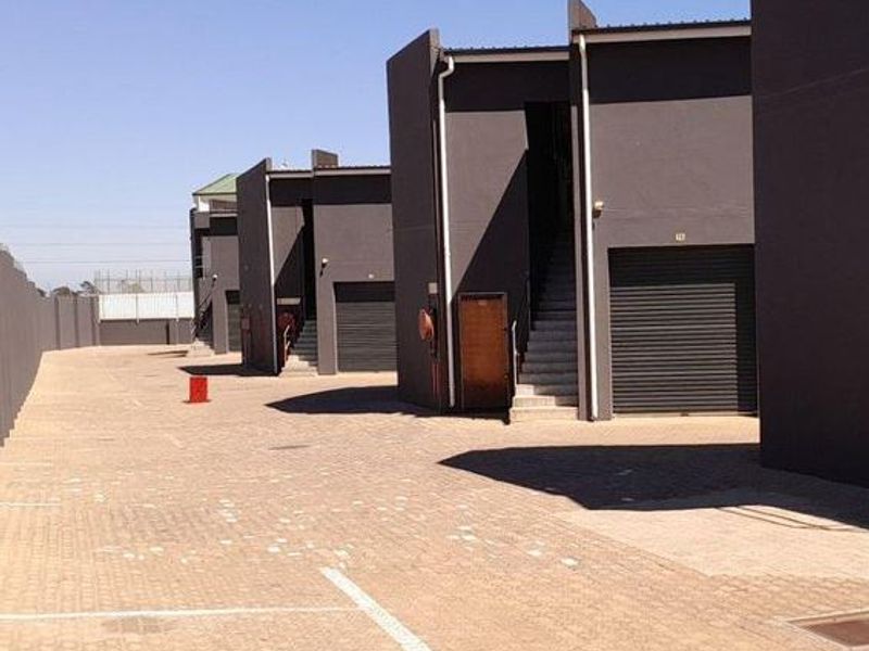 Exterior view of an industrial complex featuring multiple units with roller shutter doors, external staircases, and a paved driveway area.