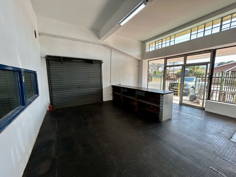 Open-plan retail or utility space featuring a built-in service counter, roller shutter door, durable rubberized flooring, and large windows providing natural light.