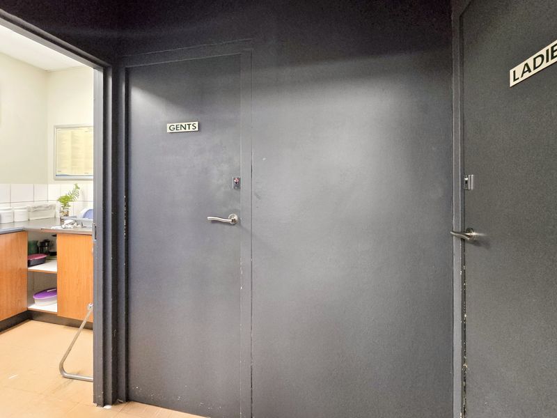 A hallway area featuring designated restroom access doors and a glimpse into a small kitchenette or utility space with built-in cabinetry.