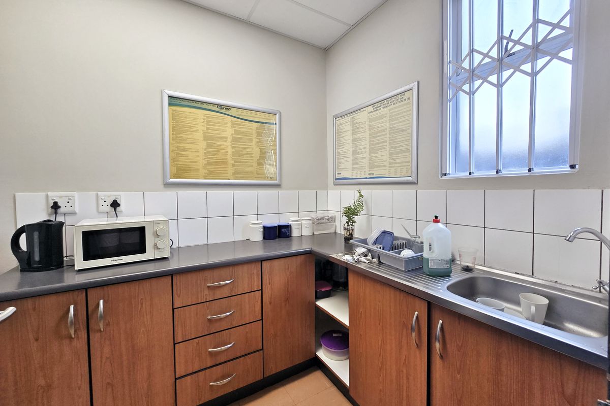 A functional office kitchenette featuring wood-finish cabinetry, laminate countertops, a stainless steel sink, and a tiled backsplash with security bars on the window.