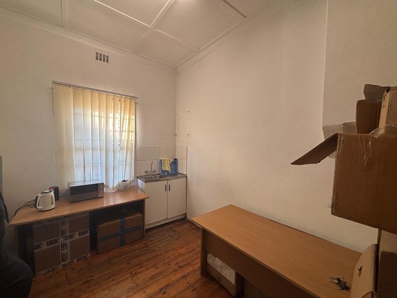 A functional office space featuring timber flooring, a small kitchenette area with sink and cabinetry, and natural light through vertical blinds.
