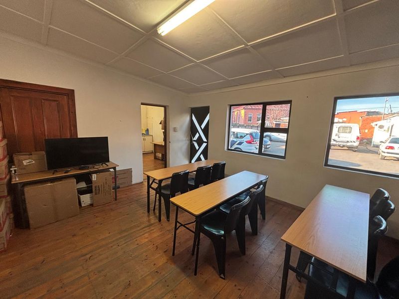 A functional office space featuring timber flooring, tiled ceiling, and natural light through aluminium-framed windows, with an adjoining utility area.