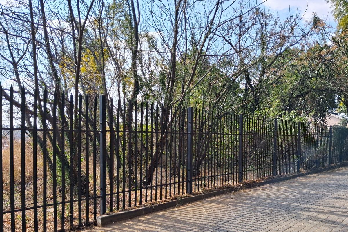 A view of the property boundary featuring a secure metal fence along a paved walkway with mature trees in the background.