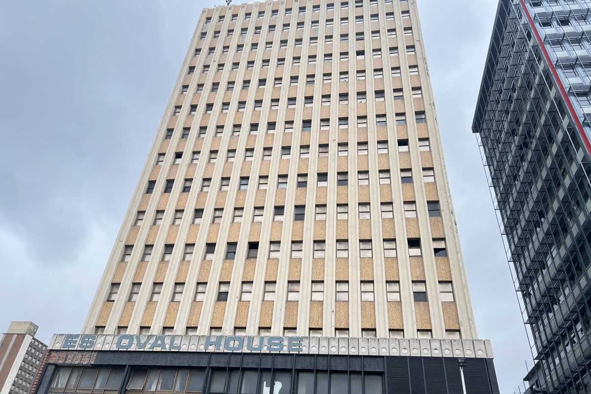 A low-angle exterior view of a multi-story apartment building facade featuring a grid window design and prominent building signage.