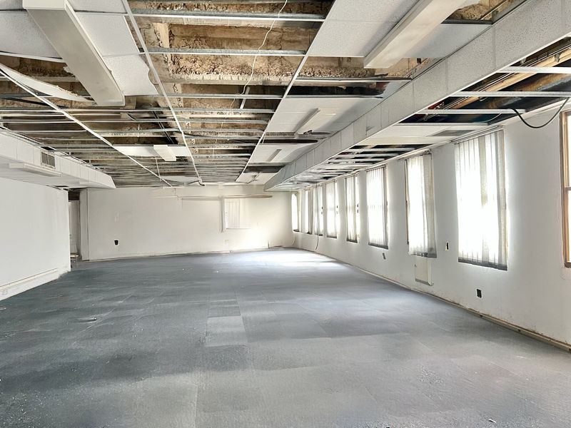 A large, vacant open-plan space featuring carpet tiles, vertical blinds, and a damaged suspended ceiling grid requiring significant repair or replacement.
