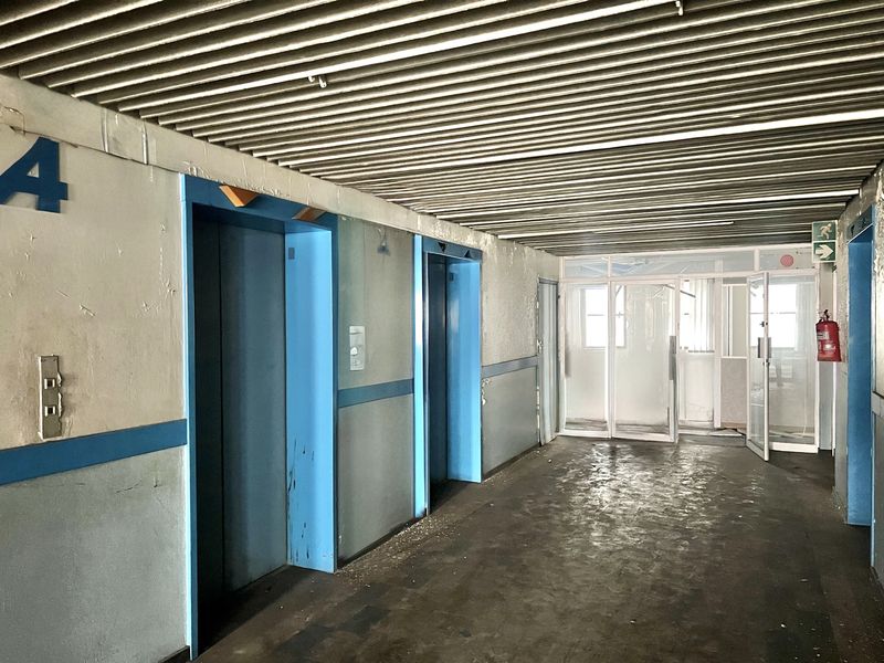 A communal apartment hallway featuring two elevator access points, a slatted metal ceiling, and glass partition doors, showing significant signs of wear and maintenance needs.