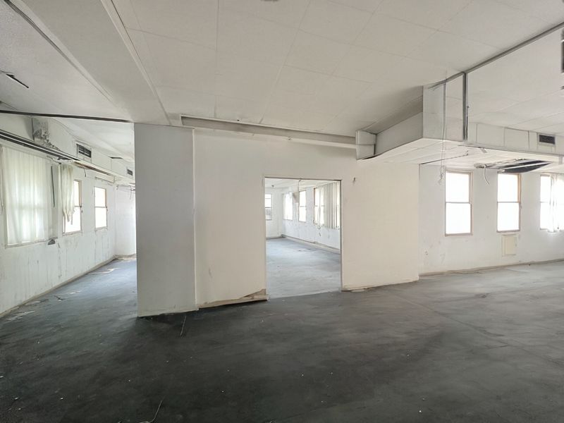 A large, vacant open-plan space featuring exposed ductwork, suspended ceiling tiles, and multiple windows, currently requiring significant renovation and cosmetic updates.