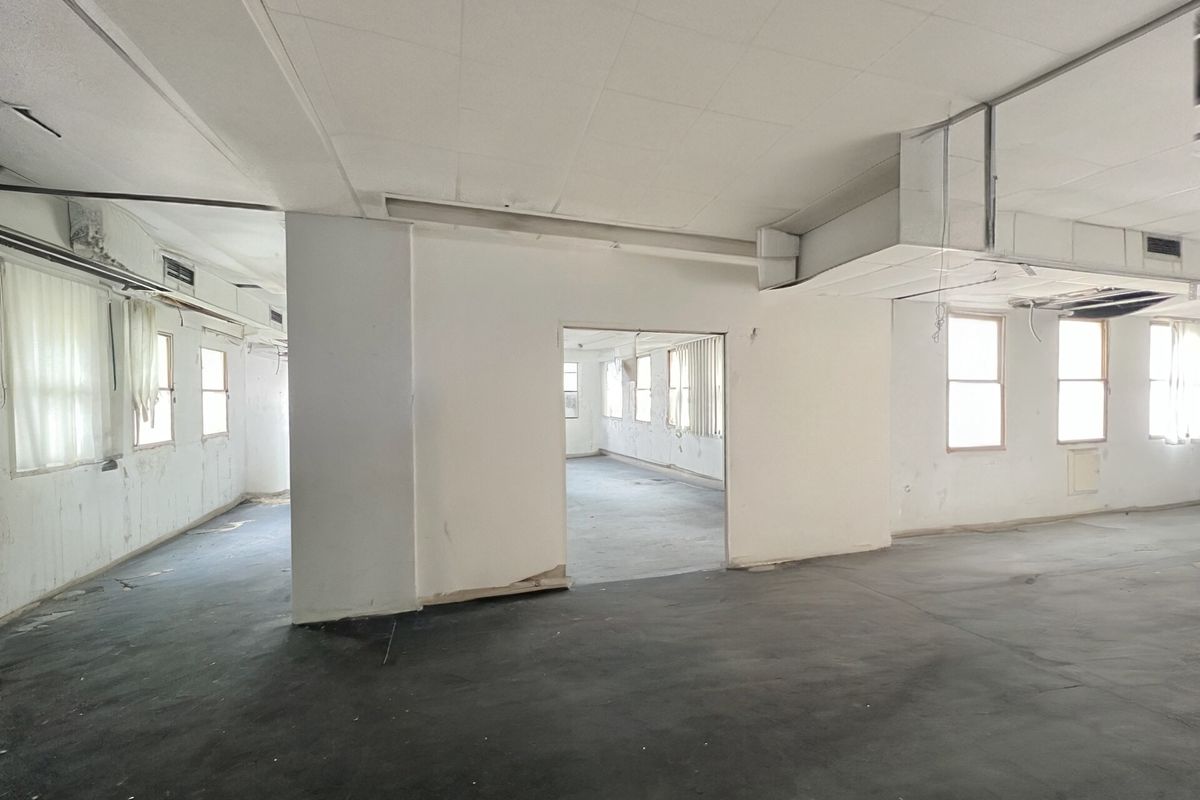 A large, vacant open-plan space featuring exposed ductwork, suspended ceiling tiles, and multiple windows, currently requiring significant renovation and cosmetic updates.
