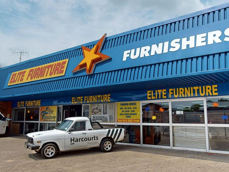 Front elevation of a commercial retail property featuring a blue corrugated metal facade, large glass storefront windows, and paved exterior parking area.