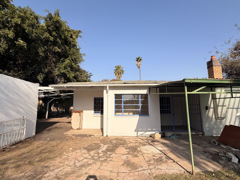 Rear exterior view of the property featuring a covered patio area, brick chimney, and paved yard space requiring maintenance.