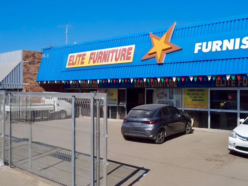 Front exterior view of a retail furniture store featuring a prominent blue metal roof, signage, and a paved parking area with security fencing.