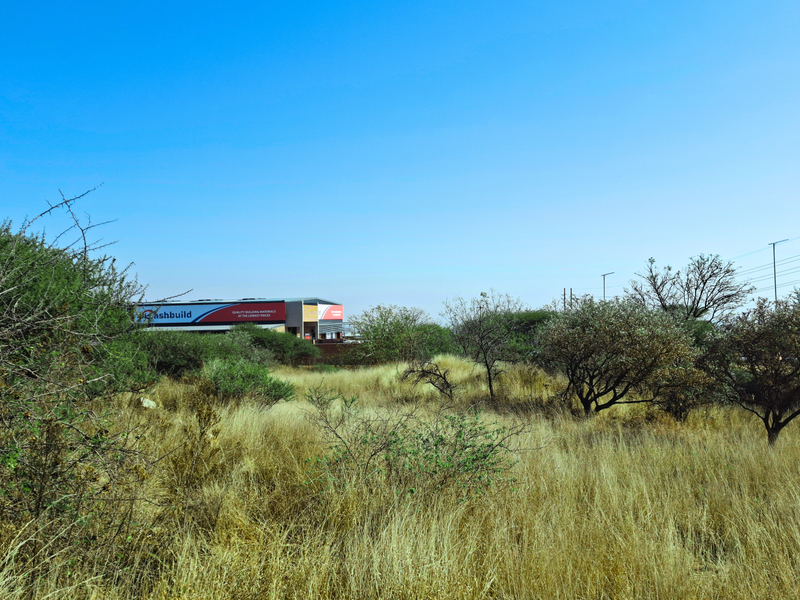 A view of an undeveloped commercial land plot featuring natural vegetation with a commercial building visible in the background.