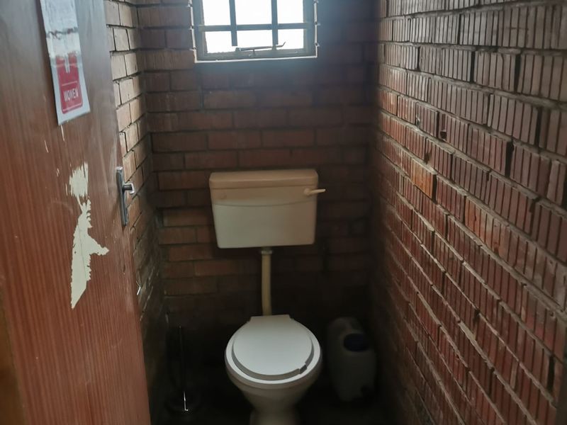 A compact industrial-style restroom featuring exposed brick walls, a single toilet, and a small window with security bars.