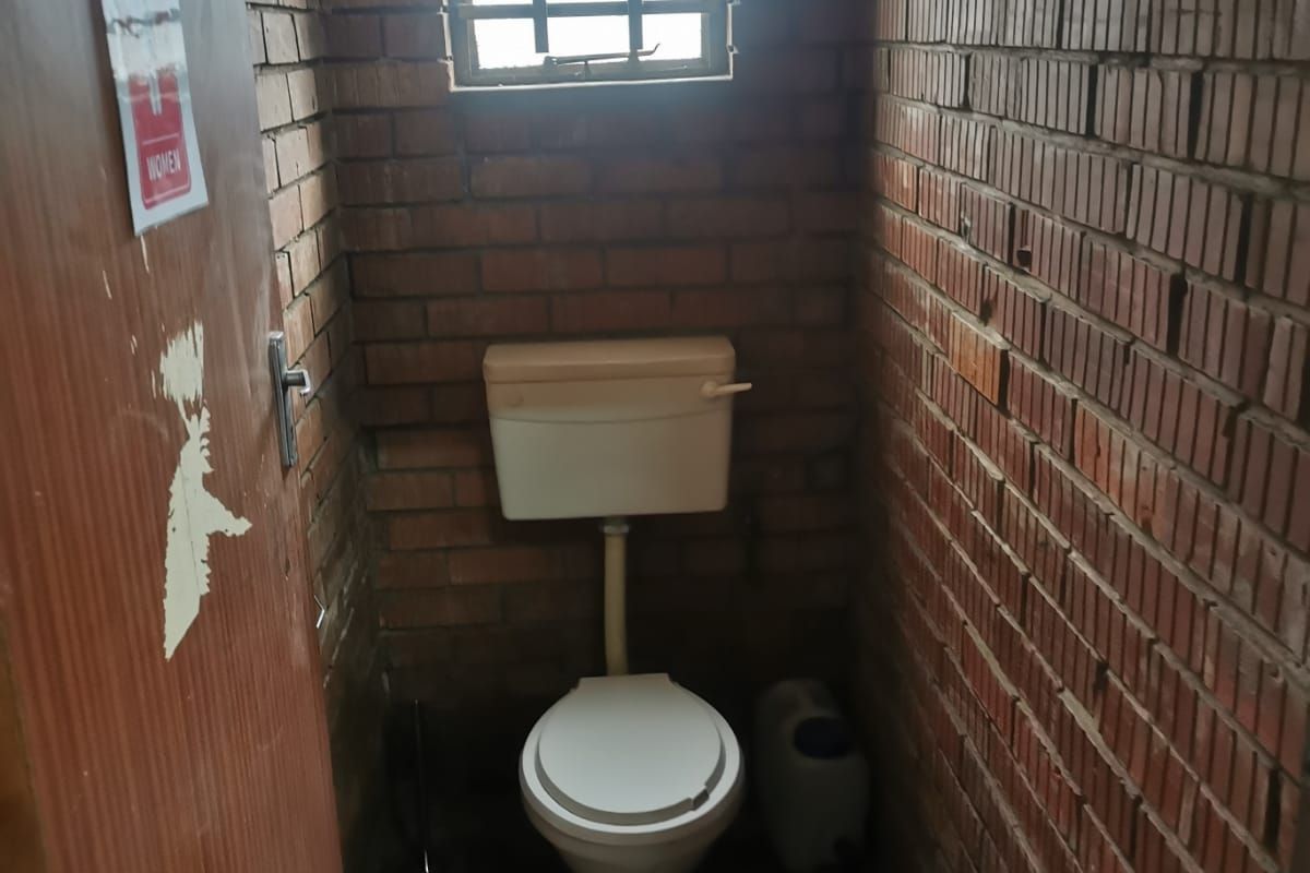 A compact industrial-style restroom featuring exposed brick walls, a single toilet, and a small window with security bars.