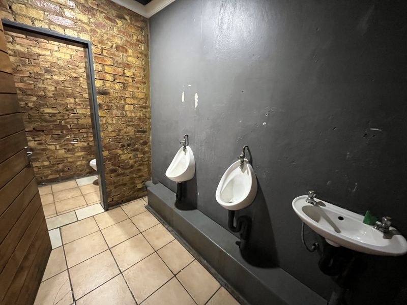 A commercial restroom facility featuring exposed brickwork, wall-mounted urinals, and a basin, requiring cosmetic updates to the wall finishes and plumbing presentation.