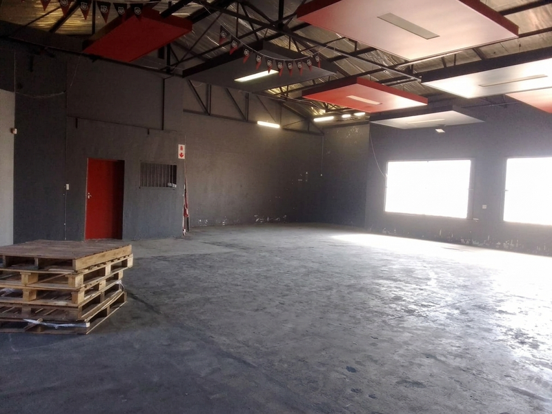 Spacious open-plan commercial area featuring exposed steel roof trusses, suspended acoustic panels, concrete flooring, and large windows providing ample natural light.