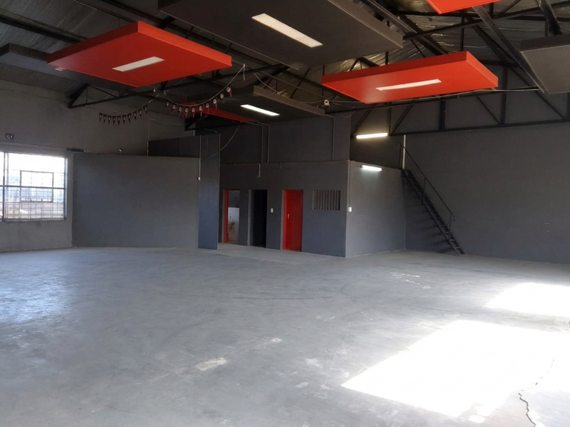 Spacious open-plan commercial warehouse area featuring exposed steel roof trusses, suspended acoustic ceiling panels, concrete flooring, and an internal metal staircase.