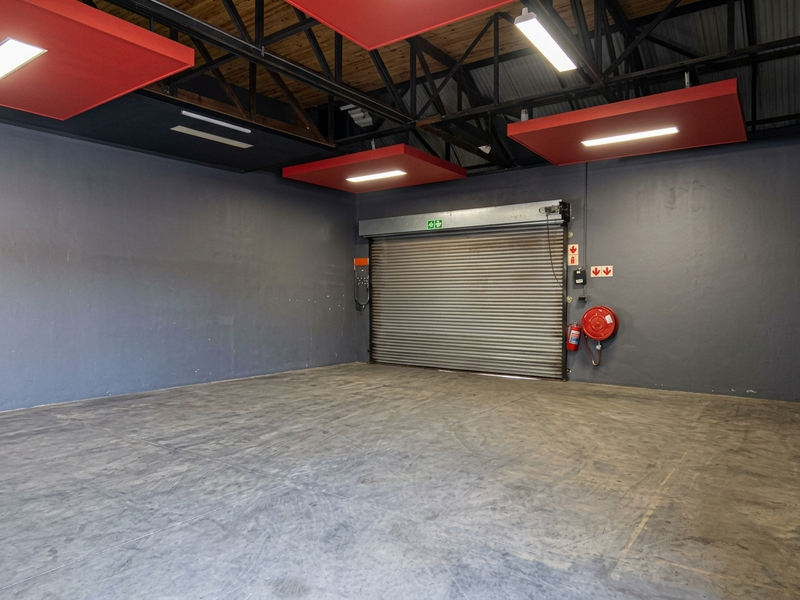 Spacious commercial garage or warehouse area featuring high exposed steel roof trusses, concrete flooring, and a secure roller shutter door.