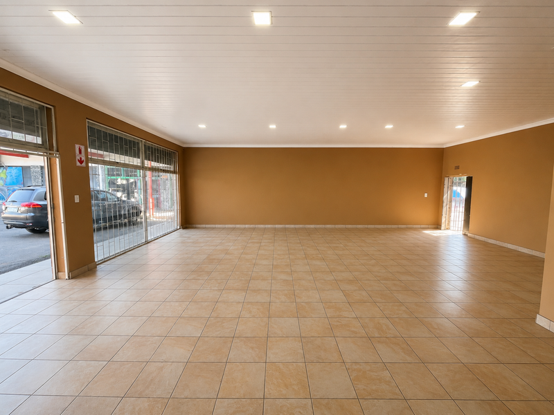 A spacious, empty retail unit featuring neutral walls, tiled flooring, recessed ceiling lighting, and a large glass storefront with security gate access.